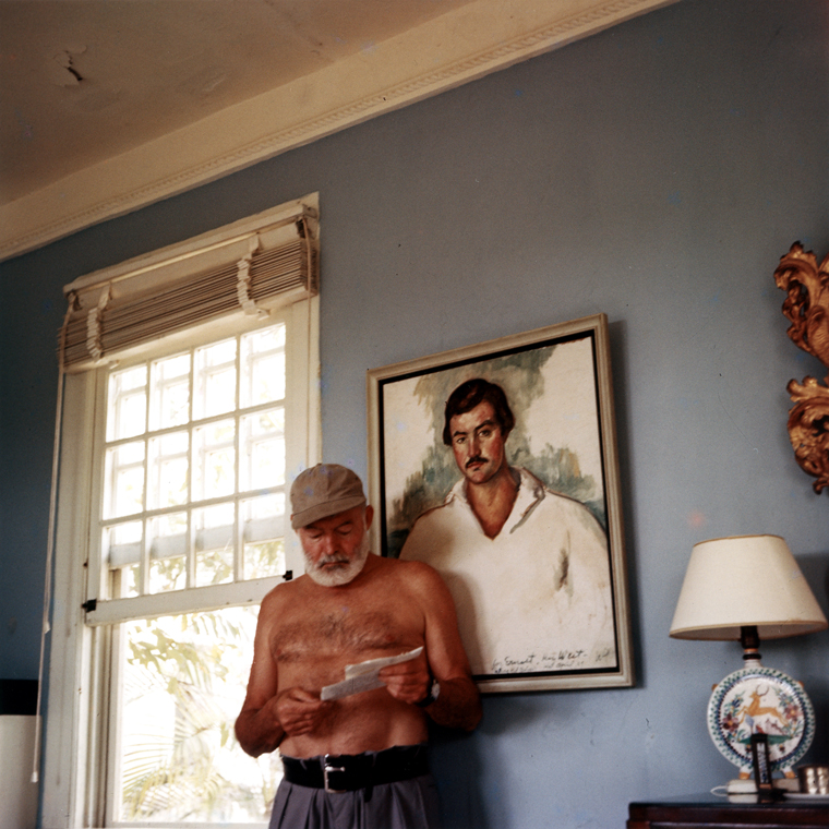 EH-C176T  Ernest Hemingway at his home in Cuba, circa 1953, standing in front of a 1929 portrait of himself by Waldo Pierce. Photograph in the Ernest Hemingway Photograph Collection, John F. Kennedy Presidential Library and Museum, Boston.